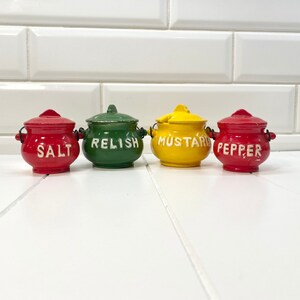 May include: Four small ceramic spice jars with lids. The jars are red, green, yellow, and red. The jars are labeled "Salt", "Relish", "Mustard", and "Pepper".