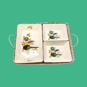Puede incluir: Un juego de servir vintage con una bandeja rectangular, un marco de metal tejido y dos platos cuadrados y uno rectangular de cerámica. Los platos tienen un fondo crema con un diseño colorido de frutas y botellas.