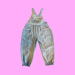 May include: Light blue acid wash denim overalls with an embroidered front pocket. The overalls have adjustable straps, elasticated cuffs, and decorative bows on the pockets. The garment is a light blue colour.