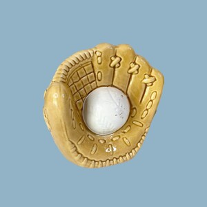 Peut inclure: Un gant de baseball en céramique beige clair tient une balle de baseball blanche. Le gant présente des coutures détaillées et des accents floraux décoratifs. Le fond est bleu clair uni.