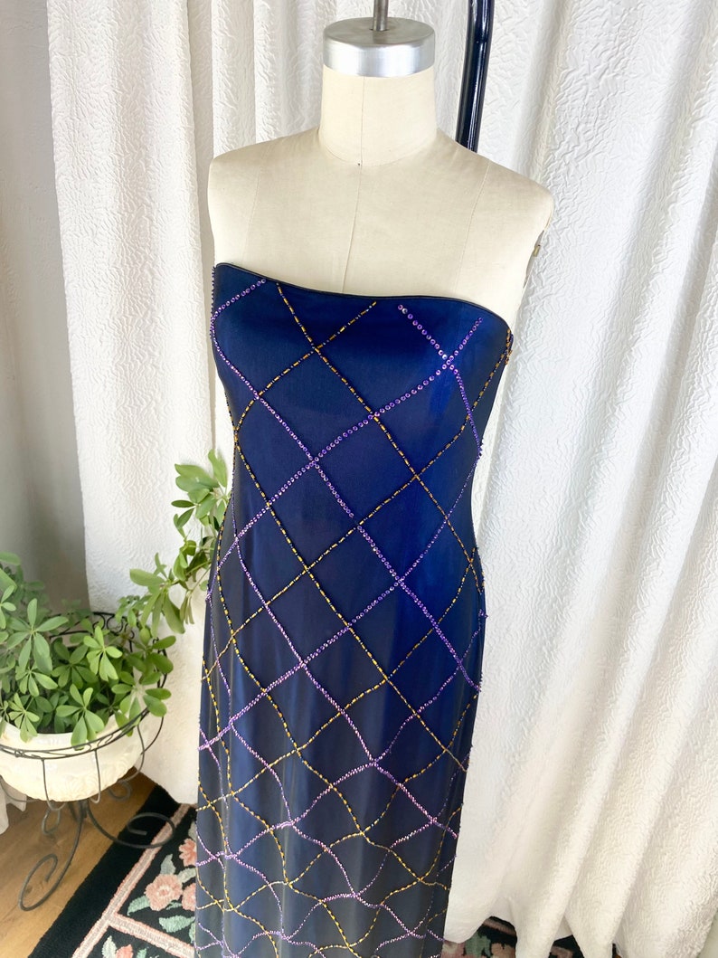 May include: A strapless, floor-length gown in a deep blue color. The gown is adorned with a geometric pattern of gold and purple beads.