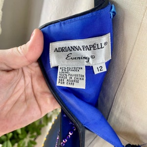 May include: A blue evening dress with black trim and a beaded design. The dress has a label that reads "Adrianna Papell Evening" and "12". The label also lists the fabric content as 97% polyester and 3% spandex. The lining is 100% polyester. The dress is made in China.