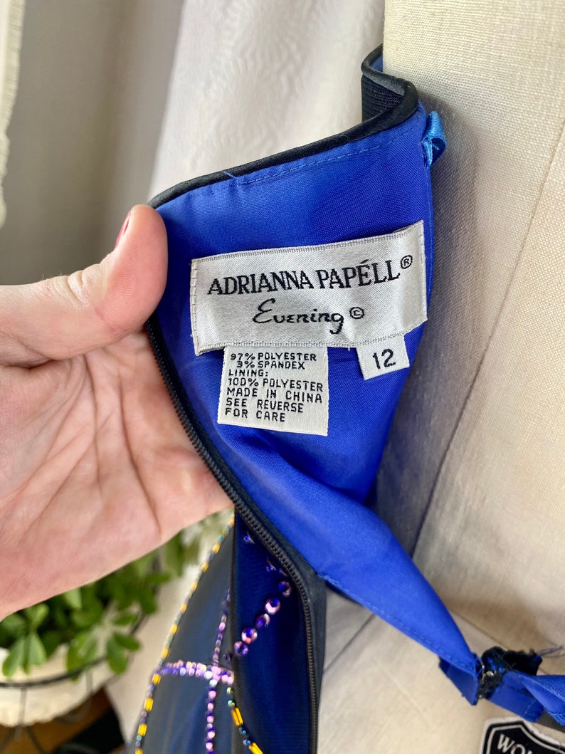 May include: A blue evening dress with black trim and a beaded design. The dress has a label that reads "Adrianna Papell Evening" and "12". The label also lists the fabric content as 97% polyester and 3% spandex. The lining is 100% polyester. The dress is made in China.
