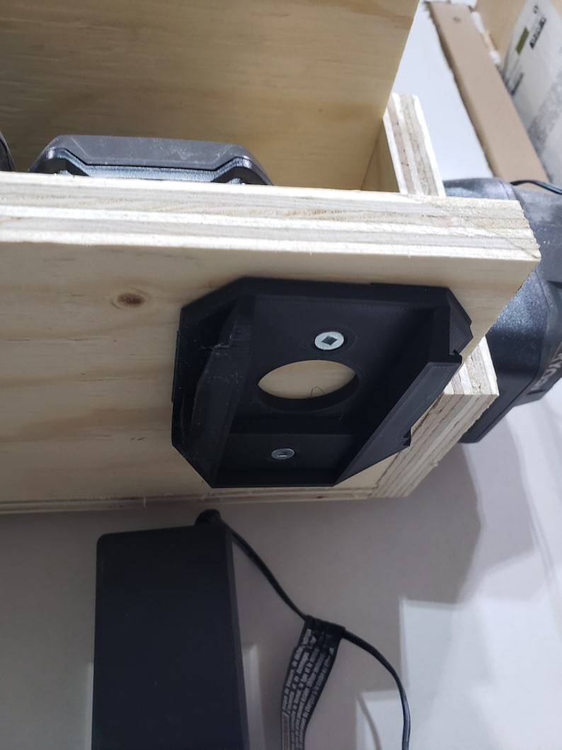 May include: A black plastic wall mount with a circular opening and two screws. The mount is attached to a wooden surface. A black power cord is visible in the foreground.