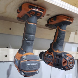 May include: Two orange and black cordless drills hanging from a wooden shelf. The drills are labeled "Brushless".