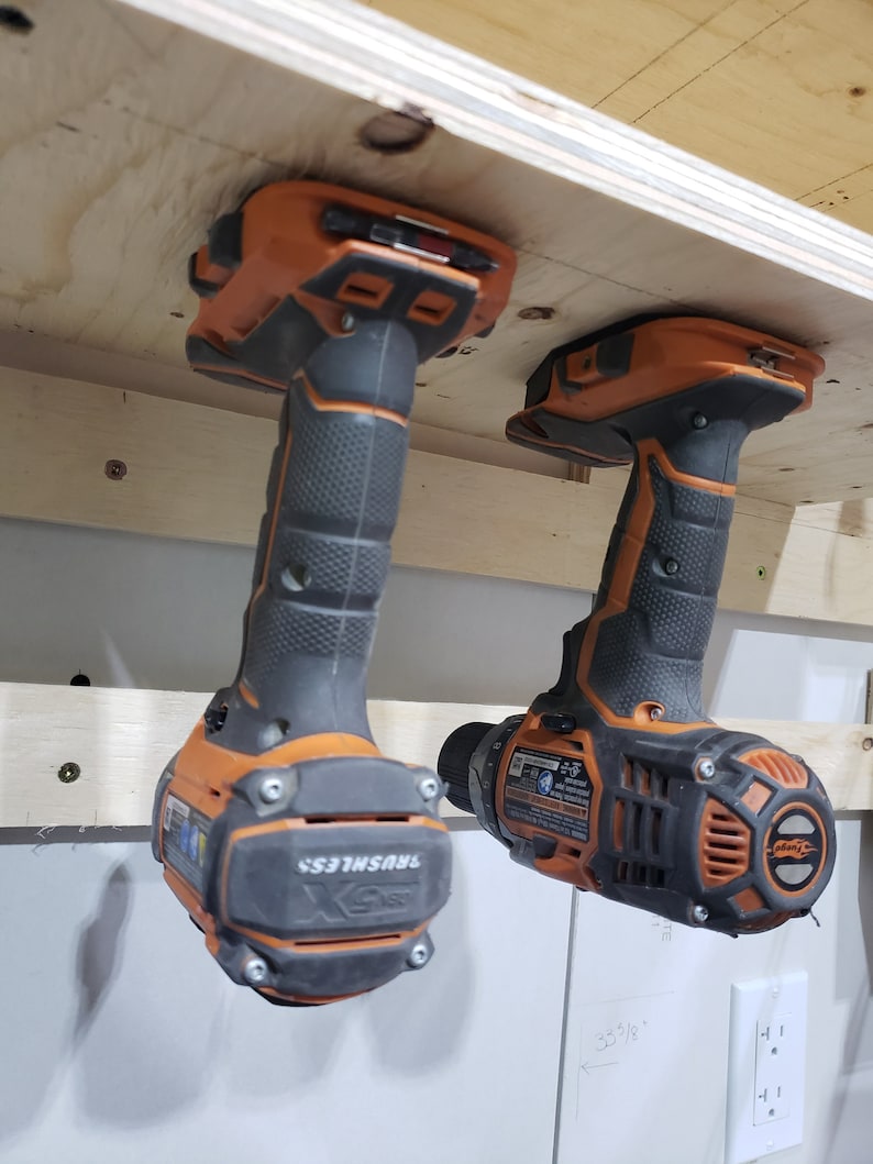 May include: Two orange and black cordless drills hanging from a wooden shelf. The drills are labeled "Brushless".