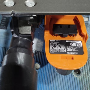 Puede incluir: Primer plano de una herramienta el&eacute;ctrica negra y naranja. La herramienta tiene un accesorio cil&iacute;ndrico negro y un compartimento para la bater&iacute;a naranja. La etiqueta del compartimento de la bater&iacute;a dice "R8657 18V". La herramienta est&aacute; sobre una superficie azul.