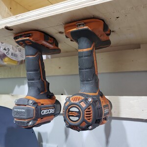 May include: Two orange and black cordless drills with the brand name "RIDGID" and the text "BRUSHLESS" are hanging from a wooden shelf.