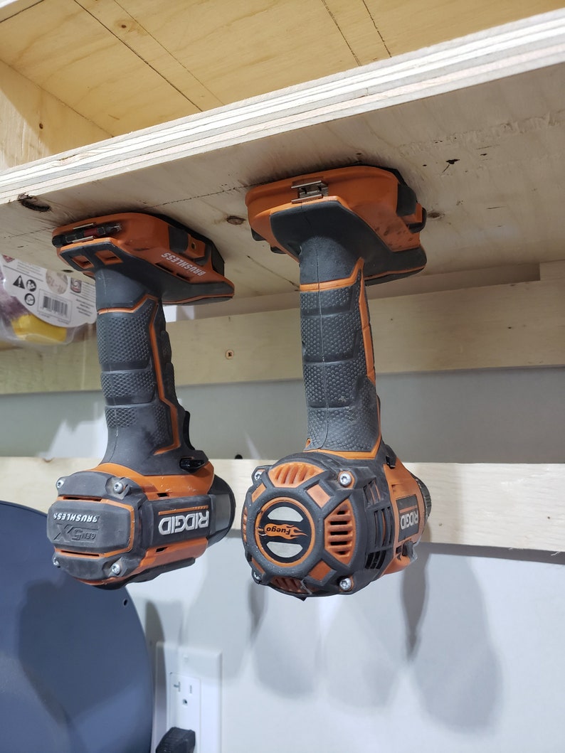 May include: Two orange and black cordless drills with the brand name "RIDGID" and the text "BRUSHLESS" are hanging from a wooden shelf.