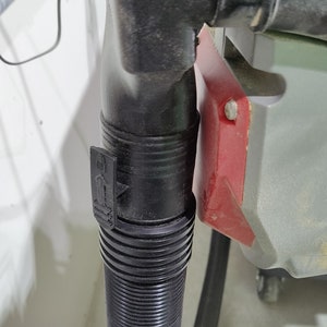 May include: Close-up of a black dust collection hose connected to a power tool. The hose has a ribbed texture and a locking mechanism with a symbol of a padlock and an arrow pointing upwards.