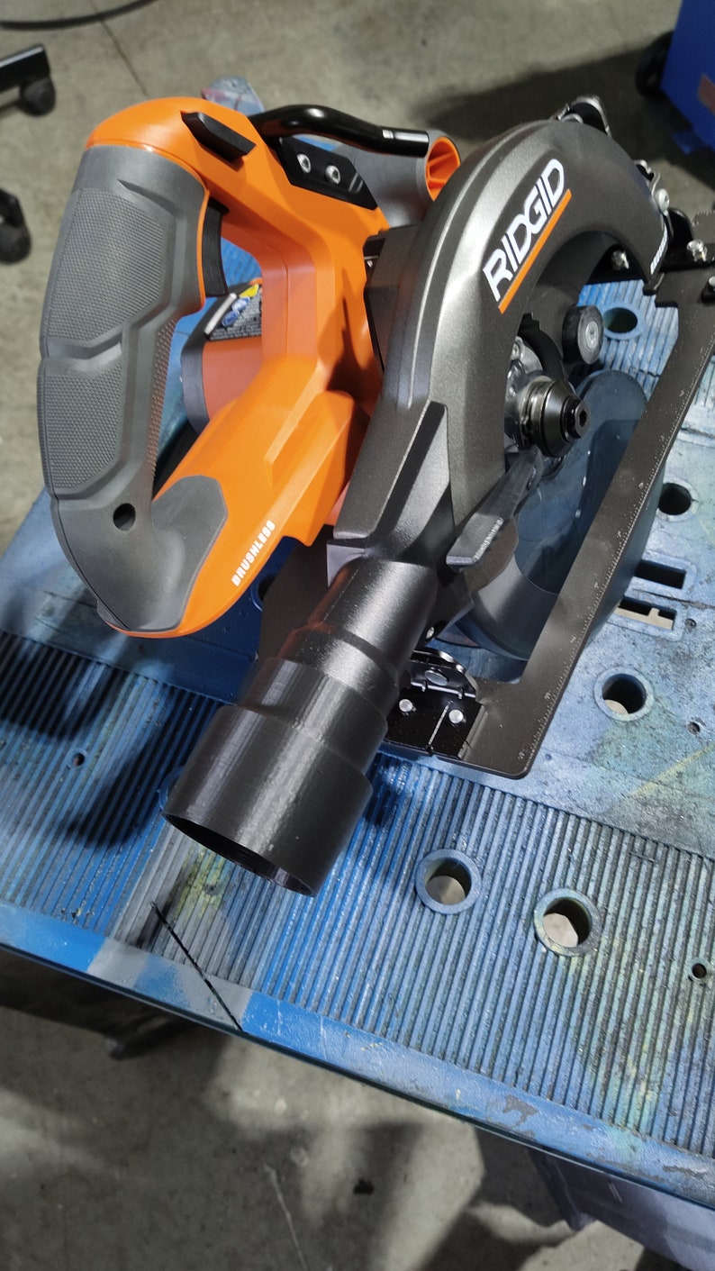 Puede incluir: Sierra circular RIDGID naranja y gris con un puerto de extracci&oacute;n de polvo negro. La sierra, adecuada para cortar diversos materiales, tiene un motor sin escobillas. Se encuentra sobre una superficie azul.