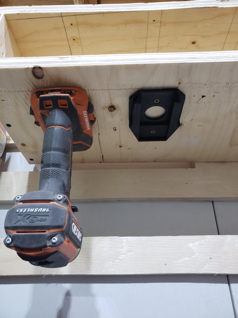 May include: A black and orange cordless drill with a brushless motor is mounted on a wooden shelf. The drill is hanging upside down with the drill bit facing up. The drill is labeled "DEWALT" and "BRUSHLESS".