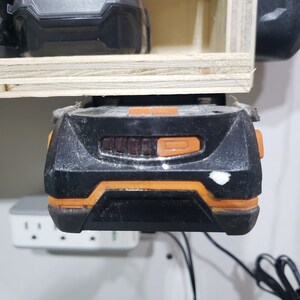 May include: A black and orange power tool battery with a charging indicator light. The battery is mounted on a wooden shelf.