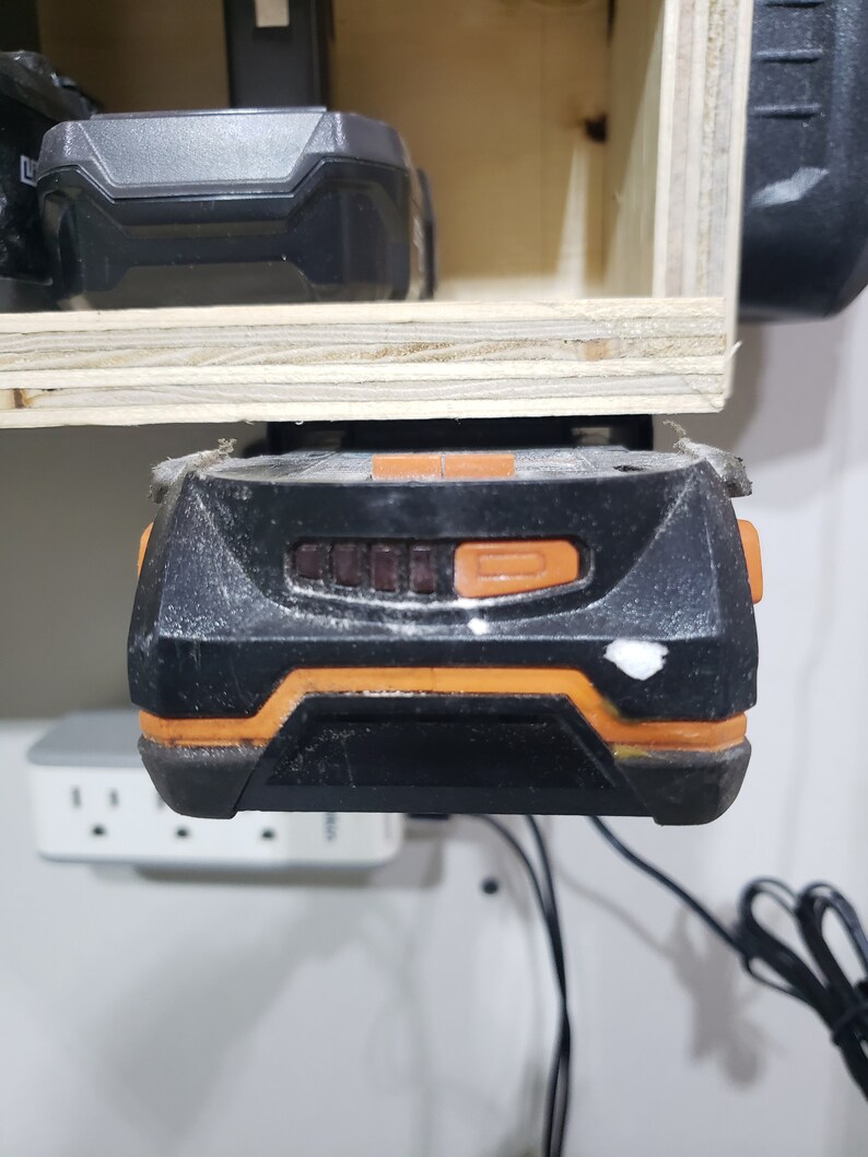 May include: A black and orange power tool battery with a charging indicator light. The battery is mounted on a wooden shelf.