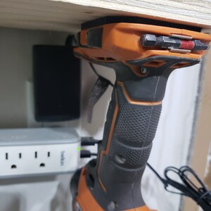 May include: An orange and black cordless drill with a black rubber grip is mounted on a wooden shelf. The drill is plugged into a white power strip with the text "Belkin" printed on it.
