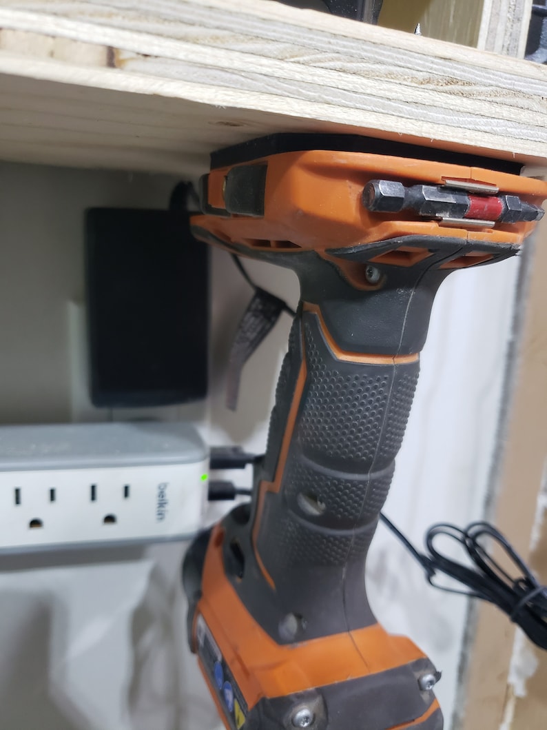 May include: An orange and black cordless drill with a black rubber grip is mounted on a wooden shelf. The drill is plugged into a white power strip with the text "Belkin" printed on it.