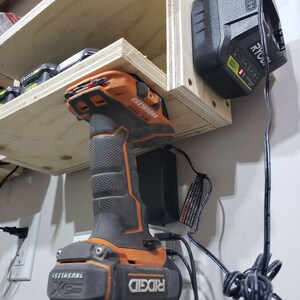 May include: A Ridgid 18V cordless drill with a brushless motor is mounted on a wall-mounted charging station. The drill is orange and black and has the text "RIDGID" and "BRUSHLESS" on it. The charging station is black and has a power cord plugged into it. A Ryobi 18V battery is also mounted on the wall-mounted charging station.
