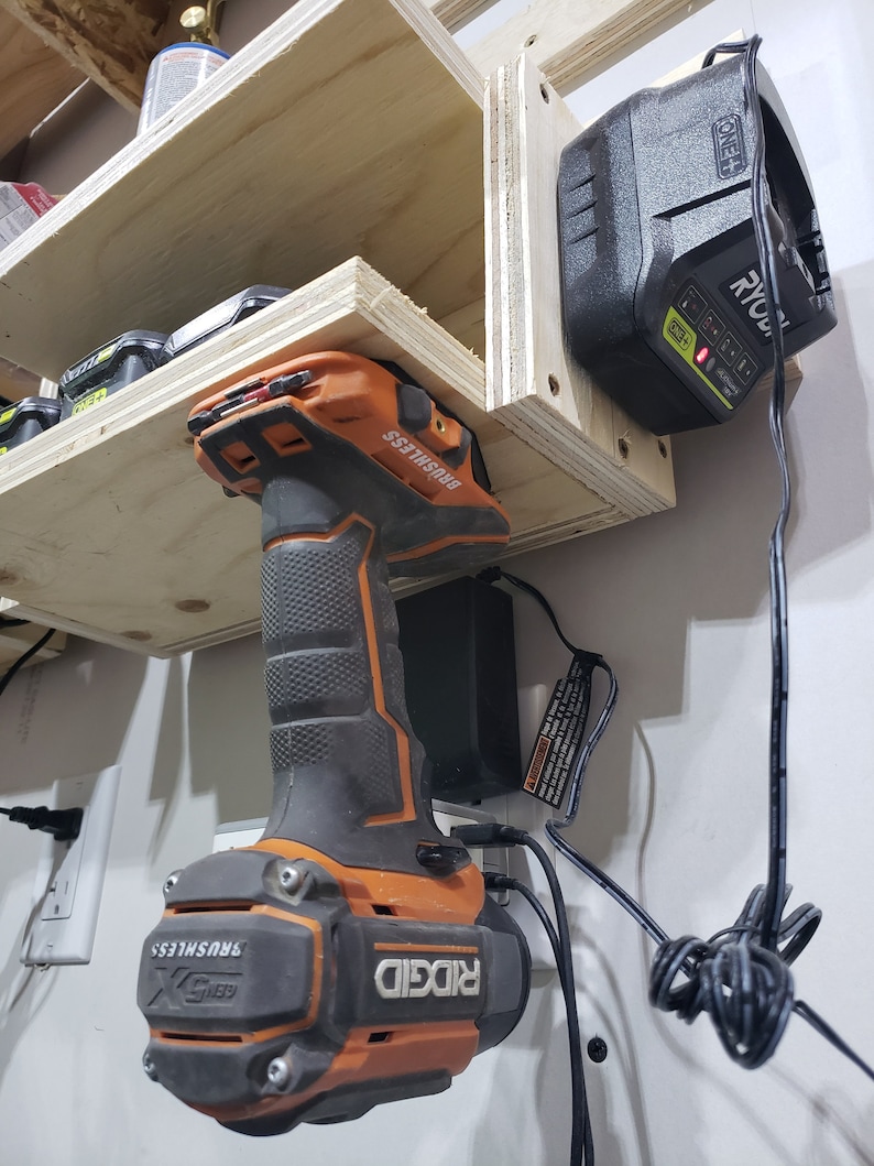 May include: A Ridgid 18V cordless drill with a brushless motor is mounted on a wall-mounted charging station. The drill is orange and black and has the text "RIDGID" and "BRUSHLESS" on it. The charging station is black and has a power cord plugged into it. A Ryobi 18V battery is also mounted on the wall-mounted charging station.