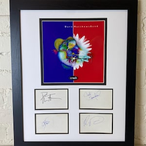 May include: A framed album cover for Dave Matthews Band's "Crash" album. The album cover features a colorful abstract design with a white flower. The frame includes four autographed cards, each with a different signature.