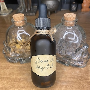 May include: A dark brown glass bottle with a dropper and a label that reads "Do as I say Oil." The bottle is tied with twine and is flanked by two skull-shaped glass bottles with cork stoppers. The background is blurred.