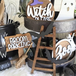 May include: A tiered tray decorated for Halloween with wooden signs that say "Witch Please", "Broom Parking", and "Boo". There is also a wooden ladder, a black bat, and a white ghost.
