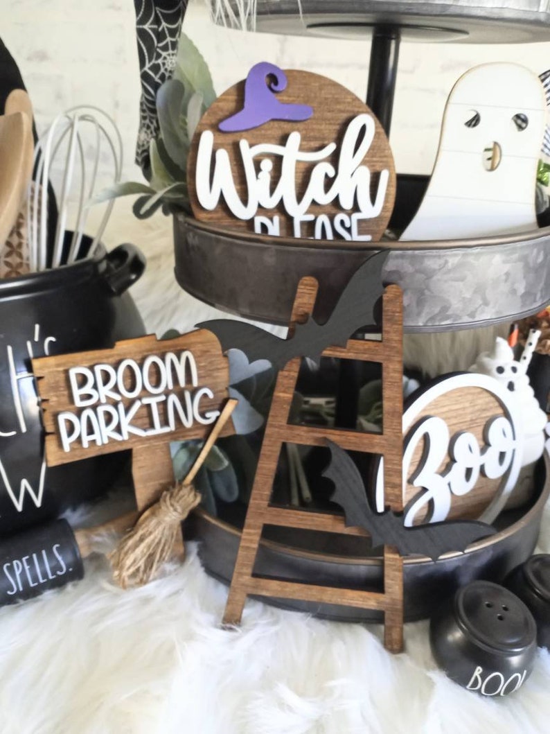 May include: A tiered tray decorated for Halloween with wooden signs that say "Witch Please", "Broom Parking", and "Boo". There is also a wooden ladder, a black bat, and a white ghost.