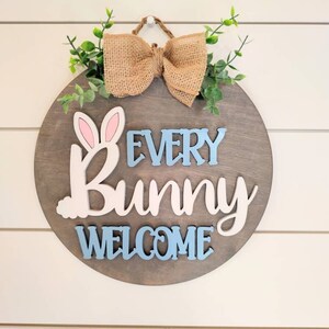 Op de afbeelding: Een rond houten bord met een strik van jute en groen. Het bord zegt "Every Bunny Welcome" in blauwe en roze letters.