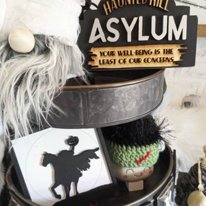 Pode incluir: Uma bandeja de tr&ecirc;s n&iacute;veis decorada para o Halloween com uma placa preta e branca que diz "Haunted Hill Asylum Your Well-Being Is The Least Of Our Concerns", uma silhueta preta e branca de um cavaleiro sem cabe&ccedil;a montando um cavalo, um gnomo de croch&ecirc; verde e branco e um esqueleto preto e branco.