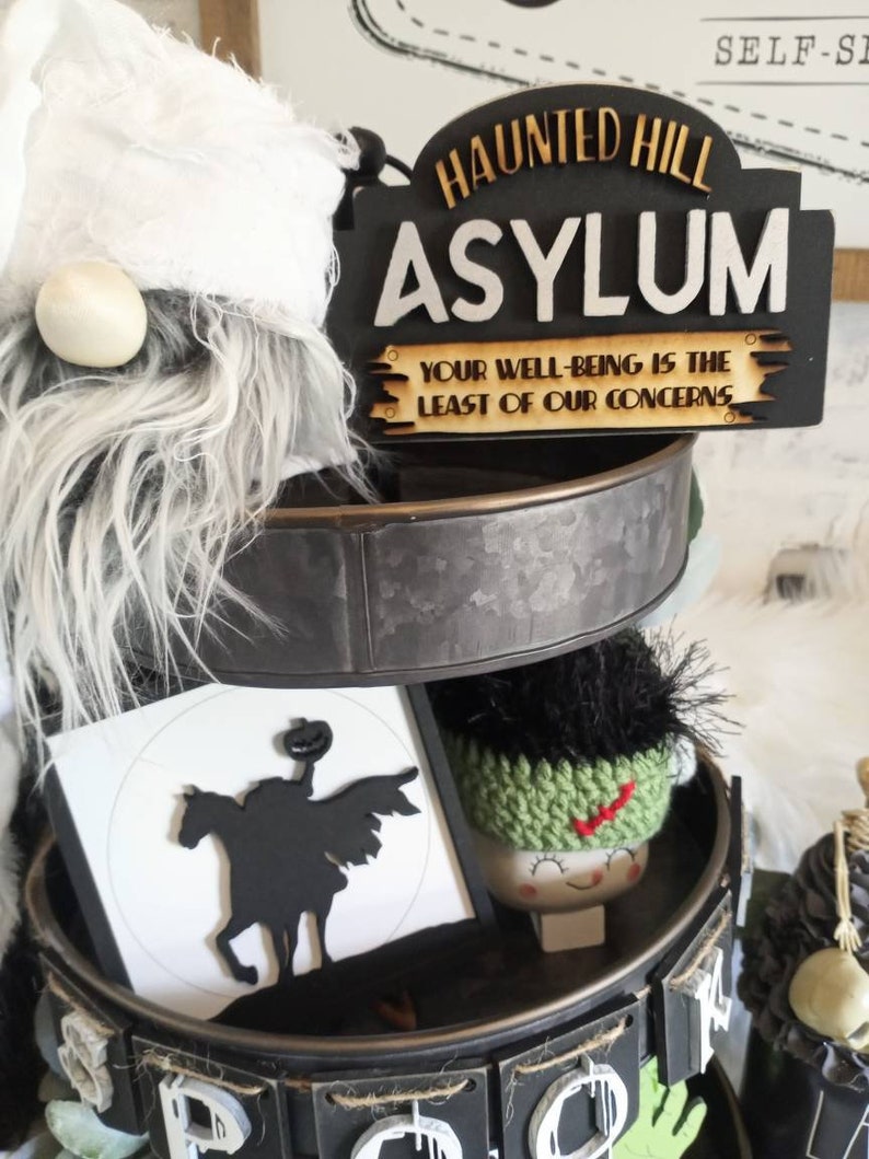Pode incluir: Uma bandeja de tr&ecirc;s n&iacute;veis decorada para o Halloween com uma placa preta e branca que diz "Haunted Hill Asylum Your Well-Being Is The Least Of Our Concerns", uma silhueta preta e branca de um cavaleiro sem cabe&ccedil;a montando um cavalo, um gnomo de croch&ecirc; verde e branco e um esqueleto preto e branco.