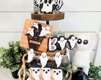May include: A three-tiered wooden tray decorated with Halloween-themed decorations. The top tier features a black and white "Spooky" sign with a ghost and spiderwebs. The middle tier has a black and white sign with a haunted house and ghosts. The bottom tier has a ghost garland and a mummy figurine.