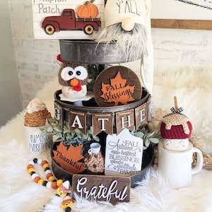 May include: A tiered tray decorated for fall with a variety of wooden signs, including "Pumpkin Patch", "Fall Y'all", "Fall Blessings", "Gather", "Pumpkin Everything", "Thankful", "Fall Autumn Harvest Blessings Splendor Gratitude Family", and "Grateful". The tray also features a gnome, a turkey figurine, a pumpkin, a coffee cup, and a candy corn garland.