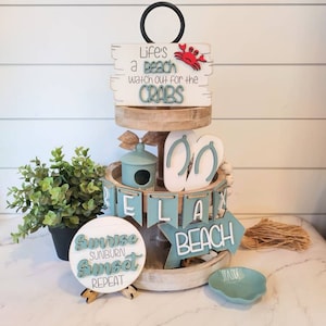 May include: A three-tiered wooden tiered tray decorated with a beach theme. The top tier features a sign that reads "Life's a beach watch out for the crabs" with a red crab. The middle tier has a banner that reads "Relax" and a pair of white flip flops. The bottom tier has a round sign that reads "Sunrise sunburn sunset repeat" and a sign that reads "Beach".