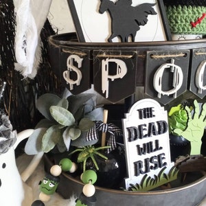 Pode incluir: Uma bandeja de metal preta de dois n&iacute;veis decorada com um tema de Halloween. A bandeja apresenta uma faixa que diz "SPOOKY", uma silhueta preta de um cavaleiro sem cabe&ccedil;a montando um cavalo, uma l&aacute;pide que diz "The Dead Will Rise", e uma guirlanda de contas verdes e pretas com um rosto de Frankenstein.