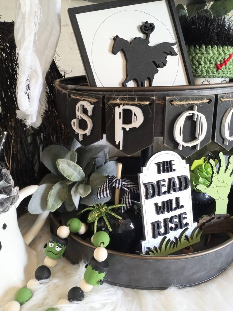 Pode incluir: Uma bandeja de metal preta de dois n&iacute;veis decorada com um tema de Halloween. A bandeja apresenta uma faixa que diz "SPOOKY", uma silhueta preta de um cavaleiro sem cabe&ccedil;a montando um cavalo, uma l&aacute;pide que diz "The Dead Will Rise", e uma guirlanda de contas verdes e pretas com um rosto de Frankenstein.