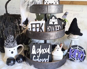 May include: A three-tiered metal tray decorated with Halloween-themed decorations. The top tier features a ghost town sign with a white ghost, the middle tier has a "EEK" sign and a "Haunted" sign with a white ghost, and the bottom tier has a "Ghoul Friend" sign with a white ghost, a white ghost-shaped mug, and a "BOO" sign with a spiderweb.