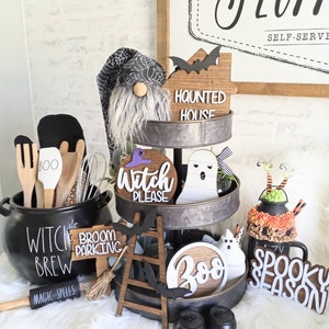 May include: A three-tiered metal stand decorated with Halloween-themed items, including a gnome, a ghost, a witch sign, a broom parking sign, a ladder, a boo sign, and a spooky season sign. The stand is on a white fluffy surface.