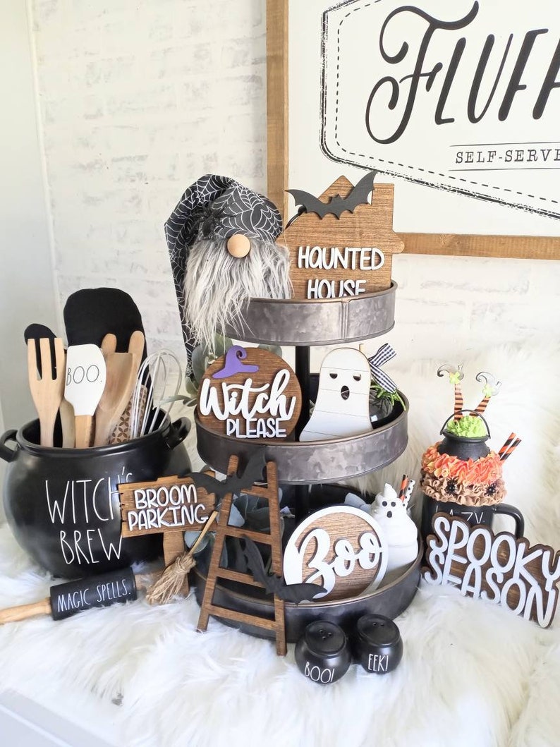 May include: A three-tiered metal stand decorated with Halloween-themed items, including a gnome, a ghost, a witch sign, a broom parking sign, a ladder, a boo sign, and a spooky season sign. The stand is on a white fluffy surface.