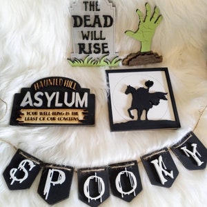 Pode incluir: Um conjunto de decora&ccedil;&otilde;es de Halloween que inclui uma placa preta e branca que diz "Haunted Hill Asylum Your Well-Being Is The Least Of Our Concerns", uma placa preta e branca com uma silhueta de um cavaleiro a cavalo, uma l&aacute;pide branca com texto preto que diz "The Dead Will Rise", uma m&atilde;o de zumbi verde e uma faixa preta e branca que diz "Spooky".
