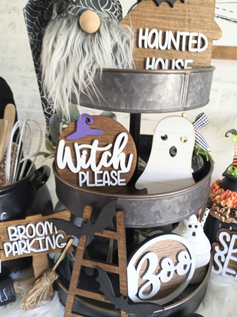 May include: A three-tiered metal stand decorated with Halloween-themed items. The top tier features a wooden sign that says "Haunted House", the middle tier has a wooden sign that says "Witch Please", and the bottom tier has a wooden sign that says "Broom Parking".