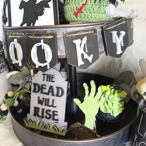 Pode incluir: Uma bandeja de tr&ecirc;s n&iacute;veis decorada para o Halloween com uma silhueta em preto e branco de um cavaleiro a cavalo, um chap&eacute;u de monstro de croch&ecirc; verde, uma faixa em preto e branco que diz "Spooky", uma l&aacute;pide que diz "The Dead Will Rise", uma m&atilde;o de zumbi verde e um cupcake preto com cobertura verde.
