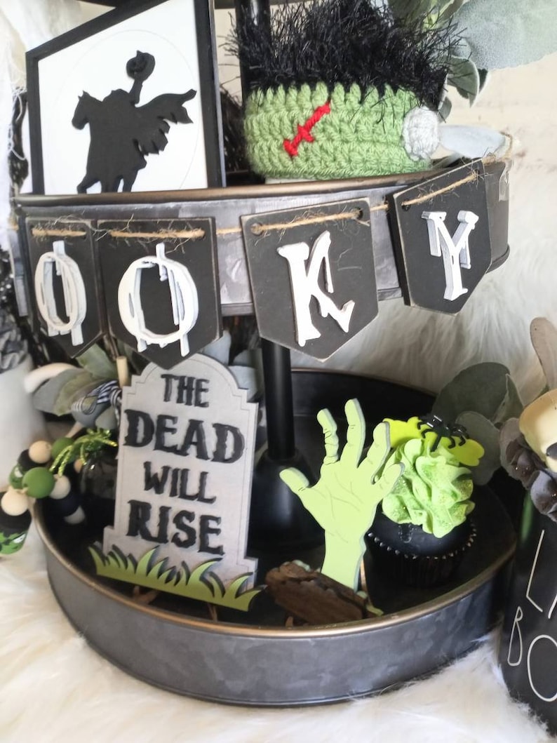 Pode incluir: Uma bandeja de tr&ecirc;s n&iacute;veis decorada para o Halloween com uma silhueta em preto e branco de um cavaleiro a cavalo, um chap&eacute;u de monstro de croch&ecirc; verde, uma faixa em preto e branco que diz "Spooky", uma l&aacute;pide que diz "The Dead Will Rise", uma m&atilde;o de zumbi verde e um cupcake preto com cobertura verde.