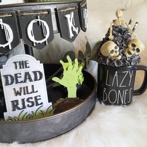 Pode incluir: Uma bandeja de tr&ecirc;s n&iacute;veis decorada para o Halloween com uma faixa preta e branca que diz "Boo", uma m&atilde;o de zumbi verde, uma l&aacute;pide que diz "The Dead Will Rise", e uma caneca preta com texto branco que diz "Lazy Bones".