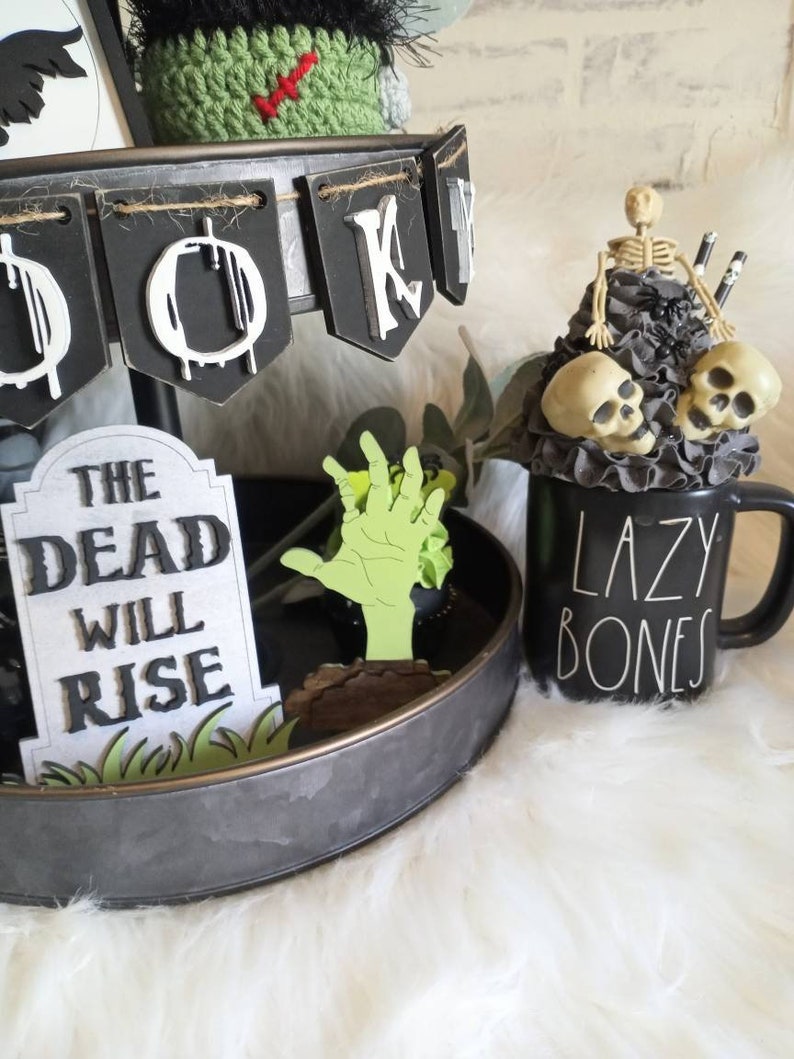 Pode incluir: Uma bandeja de tr&ecirc;s n&iacute;veis decorada para o Halloween com uma faixa preta e branca que diz "Boo", uma m&atilde;o de zumbi verde, uma l&aacute;pide que diz "The Dead Will Rise", e uma caneca preta com texto branco que diz "Lazy Bones".