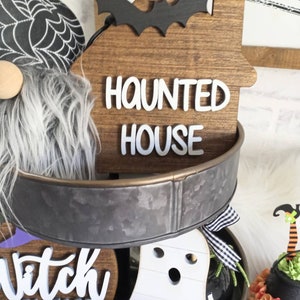 May include: A tiered metal tray decorated with Halloween themed wooden signs. The top tier features a brown wooden sign with a black bat and the text "Haunted House". The middle tier features a brown wooden sign with the text "Witch Please". The bottom tier features a white ghost cutout.