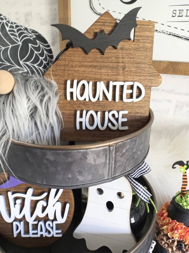 May include: A tiered metal tray decorated with Halloween themed wooden signs. The top tier features a brown wooden sign with a black bat and the text "Haunted House". The middle tier features a brown wooden sign with the text "Witch Please". The bottom tier features a white ghost cutout.
