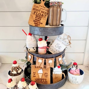 May include: A three-tiered tray decorated with a root beer float theme. The top tier features a mason jar filled with a root beer float, a wooden sign that says "Make your own root beer float", and a small potted plant. The middle tier features a small mason jar filled with a root beer float, a white sign that says "Root Beer 5¢ Floats", and a crocheted snowman. The bottom tier features a wooden sign that says "Ice Cream Shoppe", a wooden sign that says "Beat the Heat with a Frozen Treat", and a small bowl filled with a root beer float. The tray is decorated with a banner of wooden ice cream cones.