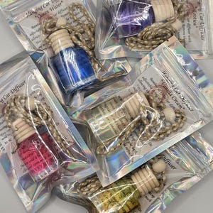 May include: Several hanging car diffusers in clear plastic bags. Each diffuser has a wooden top, a glass bottle with colored liquid, and a decorative string. The labels read "Ocean Water", "Erotica", and "Vanilla Cream".