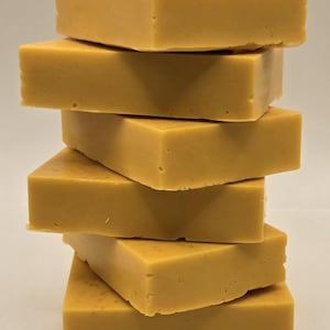 May include: A vertical stack of six rectangular bars of solid mustard-yellow soap. Each bar has a smooth, matte finish with subtle, tiny dark brown speckles distributed throughout the surface, creating a uniform appearance across the entire stack.