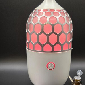 May include: White and silver essential oil diffuser with a honeycomb design and a red glow. A small bottle of jasmine oil and a note are next to the diffuser. The diffuser has a power button.
