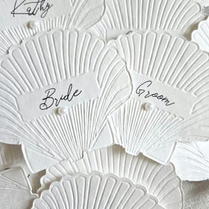 May include: Close-up of white seashell-shaped place cards with embossed details. Each card features a label with handwritten names or titles such as "Bride" and "Groom". Small pearl accents are visible.